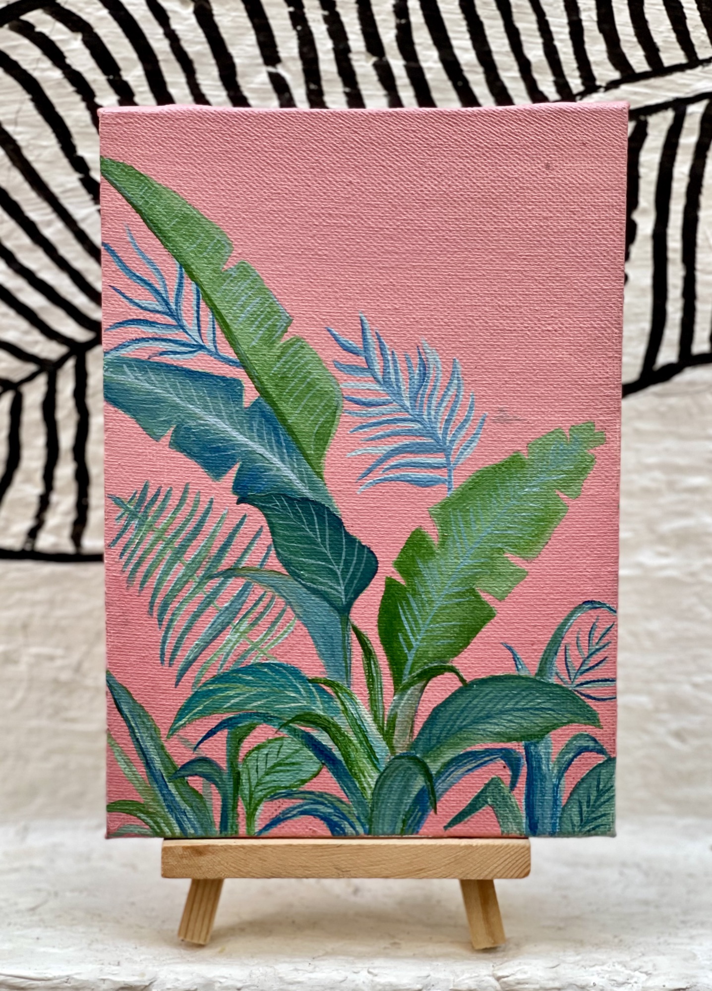 Small acrylic painting of banana leaves and palm fronds in green and blue on a pink background, propped on a mini easel