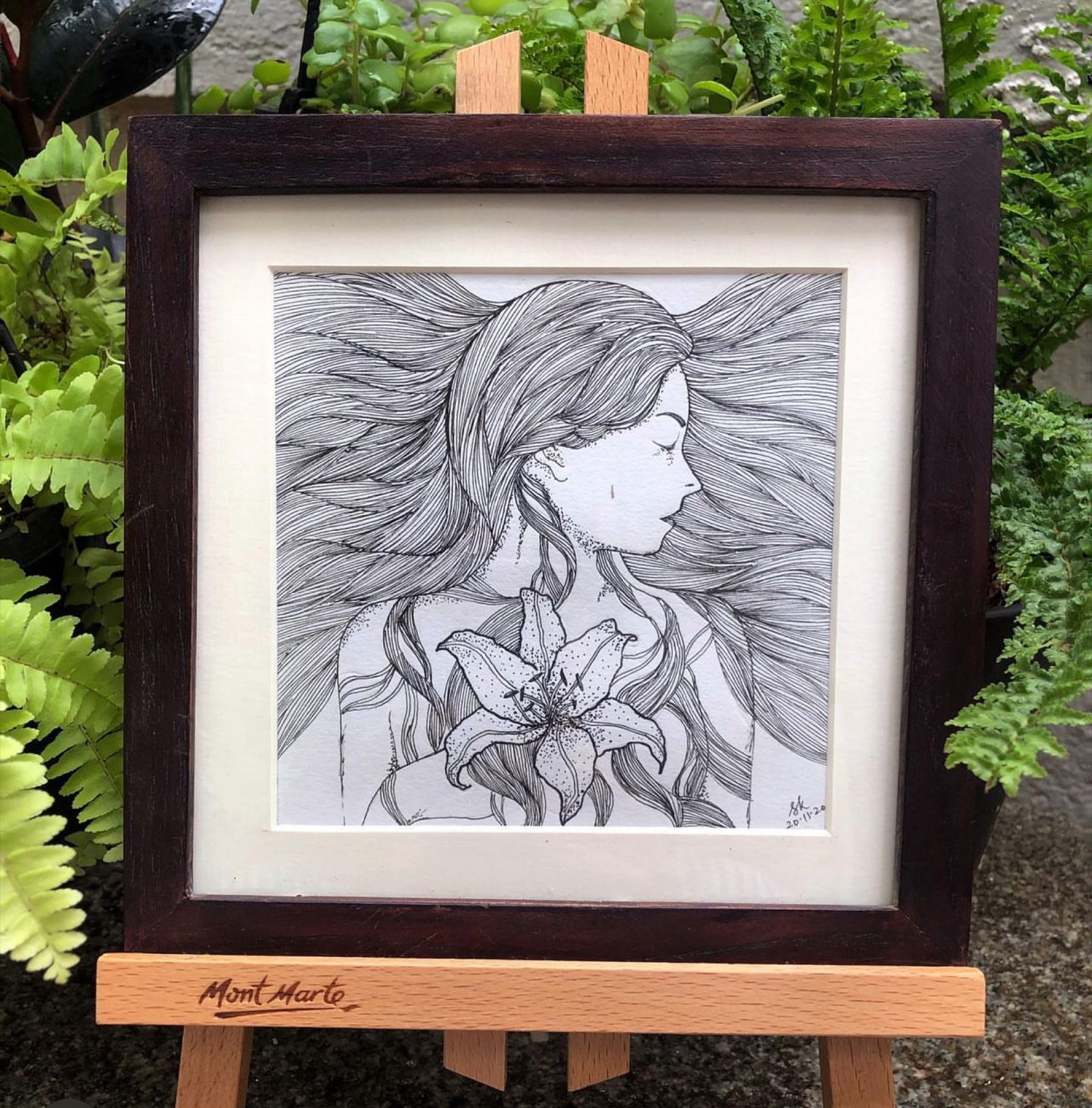 Framed black-and-white pen illustration of a woman's profile with flowing hair and a lily, displayed on an easel among real ferns